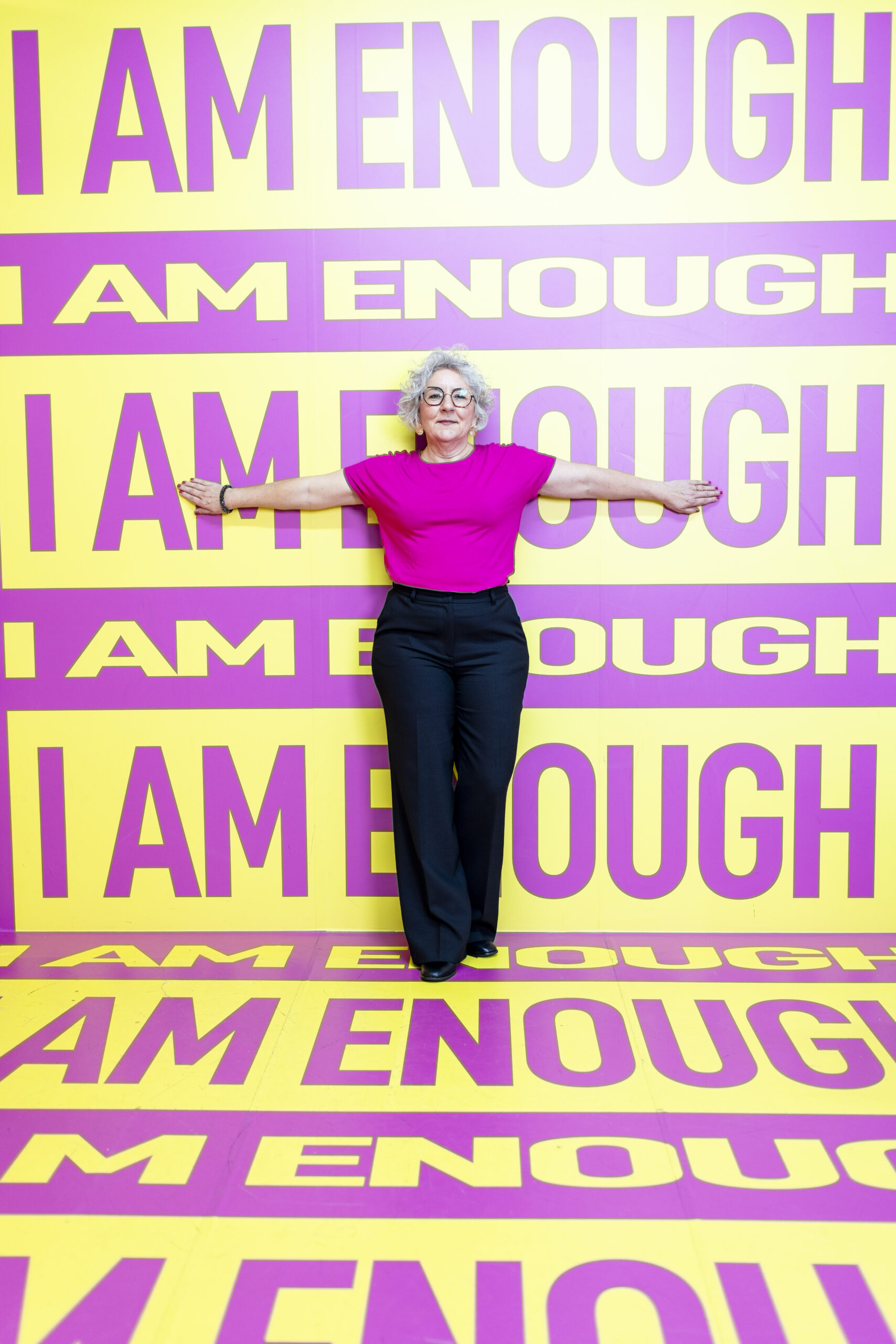 Authentisches Personal-Branding-Foto einer Frau in pinkem Oberteil steht mit ausgebreiteten Armen vor einer gelb-pinken Wand mit der Aufschrift „I AM ENOUGH“. Das authentische Personal-Branding-Foto vermittelt Selbstbewusstsein, Selbstwert und eine starke persönliche Markenpräsenz.