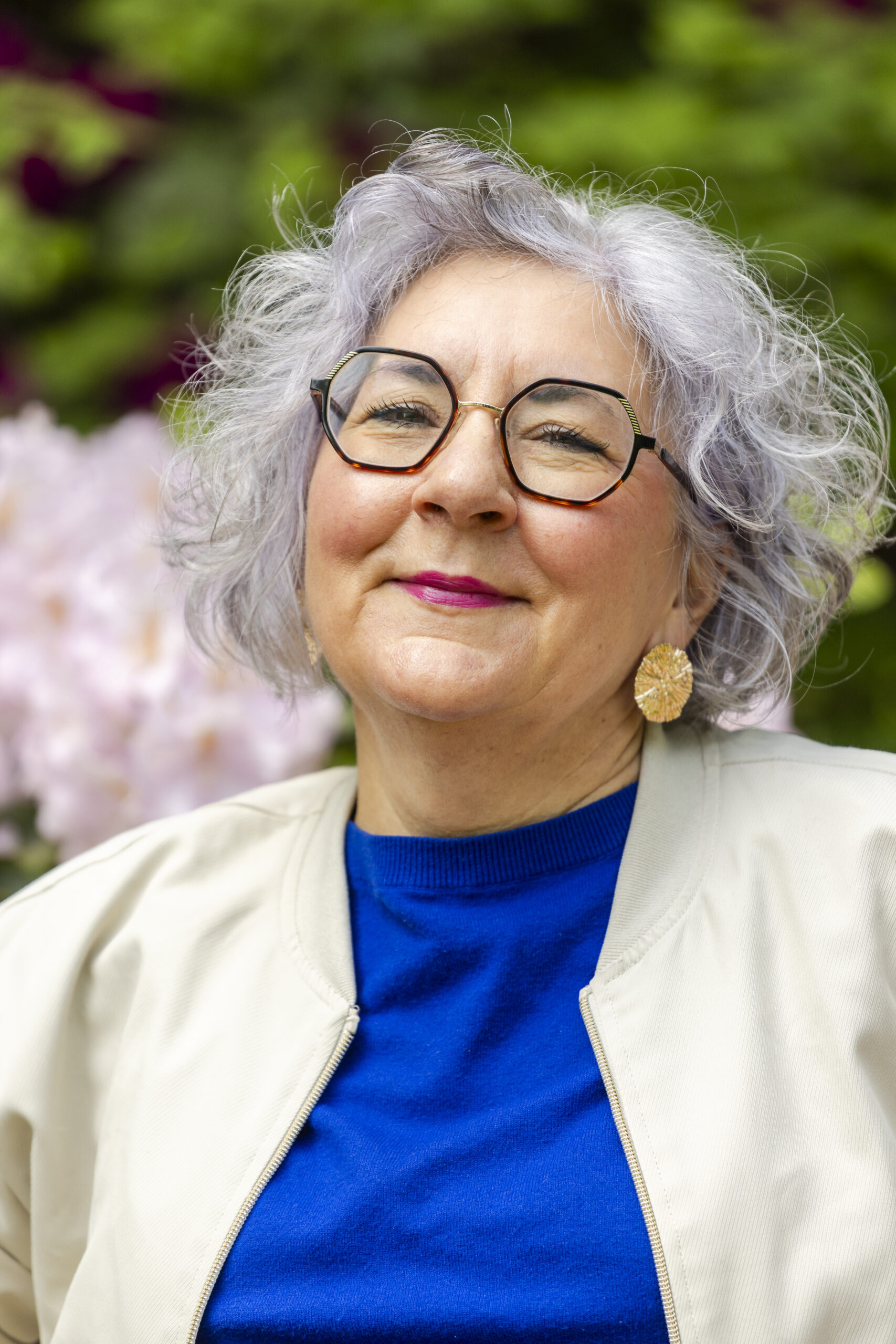 Authentisches Personal-Branding-Nahporträt einer lächelnden Frau mit silbergrauen lockigen Haaren und Brille vor einem blühenden Garten. Authentisches Personal-Branding-Foto mit natürlichem Hintergrund für eine persönliche und professionelle Markenpräsenz.