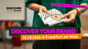 Eine Person hält mehrere Flyer des BarCamps mit einem stilisierten Auge und dem Text „DISCOVER YOUR BRAND“ in der Hand. Darüber liegt ein pinkes Label mit schwarzer Schrift: „offline BarCamp in Frankfurt am Main“. Darunter steht in großer Schrift: „DISCOVER YOUR BRAND“ und in orange hinterlegt: „16.10.2026 in Frankfurt am Main.“ Im Hintergrund ist unscharf ein Präsentationsbildschirm zu sehen. Ein BarCamp zum Thema Sichtbarkeit.