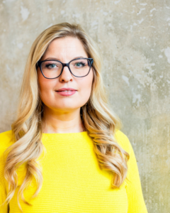 Eine Frau mit langen blonden Haaren, schwarzer Brille und gelbem Oberteil blickt direkt in die Kamera. Sie steht vor einer hellen, strukturierten Wand. Das Bild wird im Kontext von Personal Branding und der Beratung DISCOVER YOUR BRAND von Ann Sophie Detje verwendet.