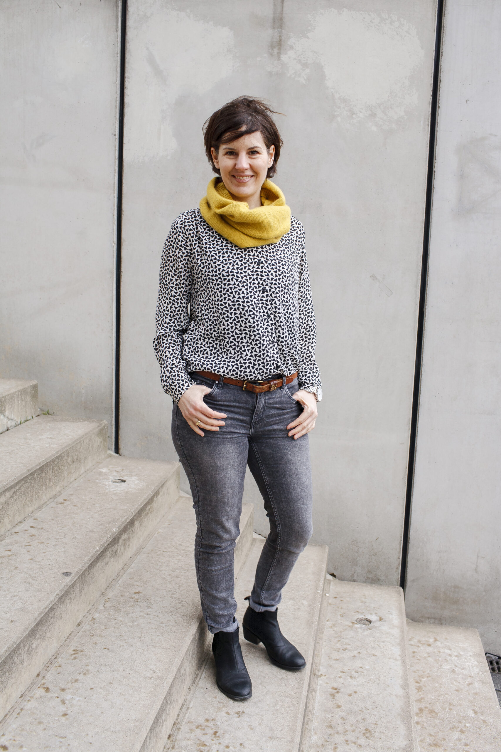 Eine lächelnde Frau mit kurzen dunklen Haaren steht auf einer Betontreppe vor einer grauen Betonwand. Sie trägt einen gemusterten Pullover, einen gelben Schal, Jeans und schwarze Stiefeletten. Das Bild wird im Kontext von Personal Branding und der Beratung DISCOVER YOUR BRAND von Ann Sophie Detje verwendet.